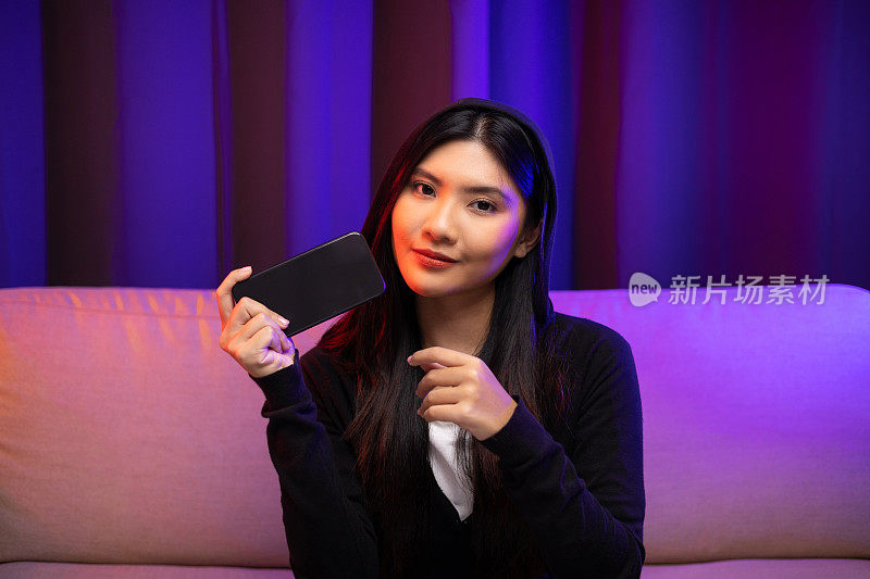 用手机玩游戏。年轻的亚洲漂亮女人坐在沙发上拿着智能手机在客厅。快乐女性Pro Streamer中国人穿着连帽衫玩游戏与霓虹灯。
